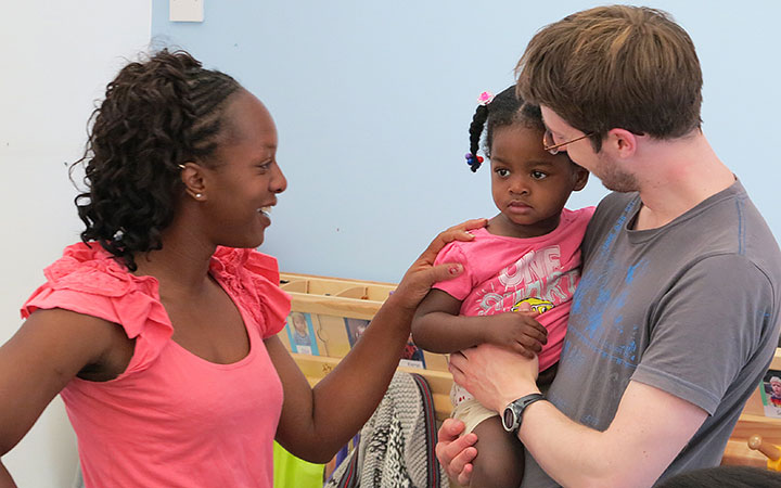 A mother supports her young daughter in a typical transition in childcare as she is welcomed by a nursery teacher