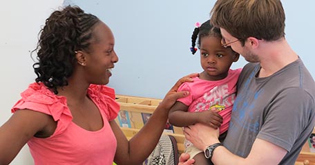 A mother supports her young daughter in a typical transition in childcare as she is welcomed by a nursery teacher