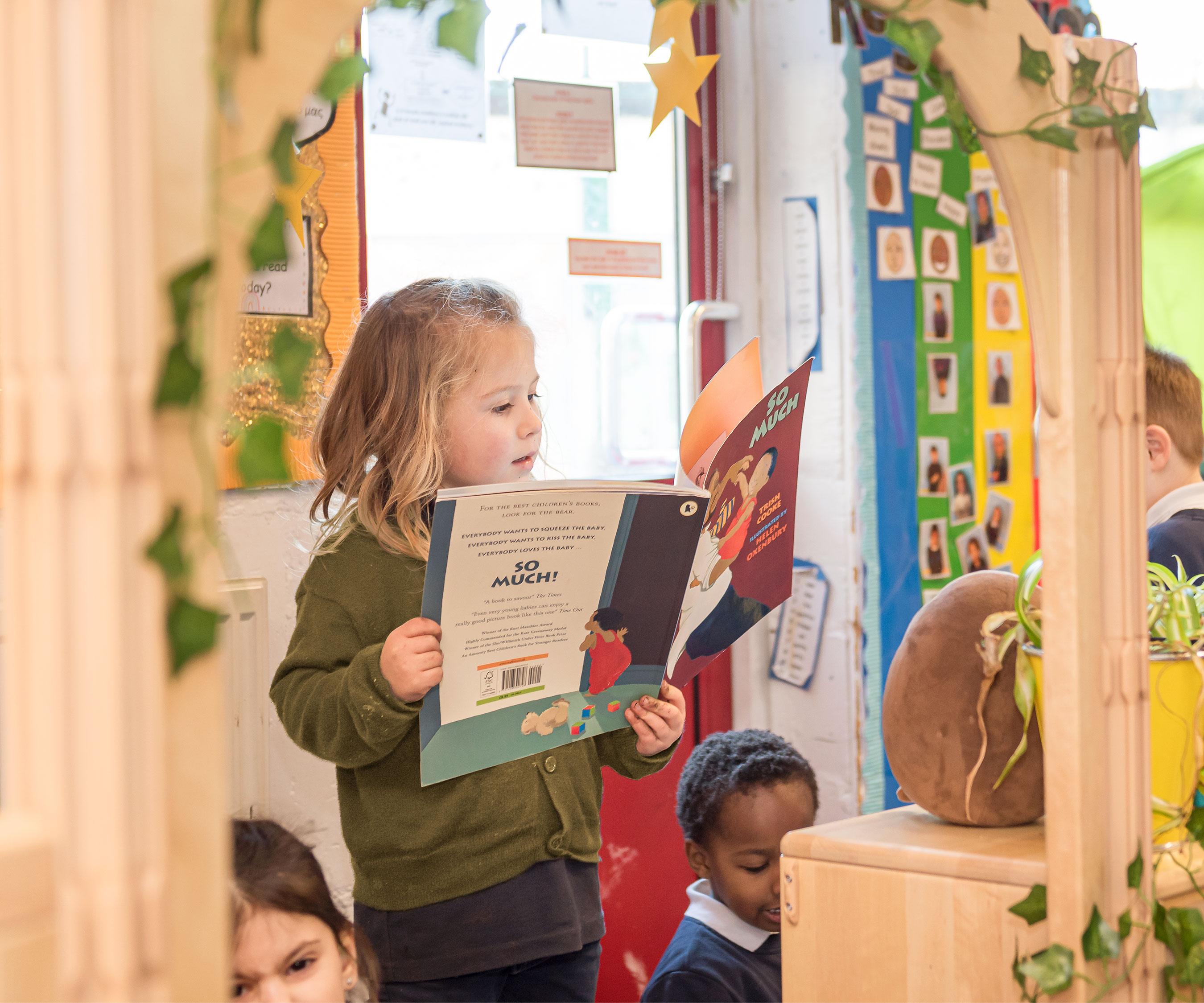 A young child with blonde hair stands, engrossed in a picture book, framed by a wooden archway.