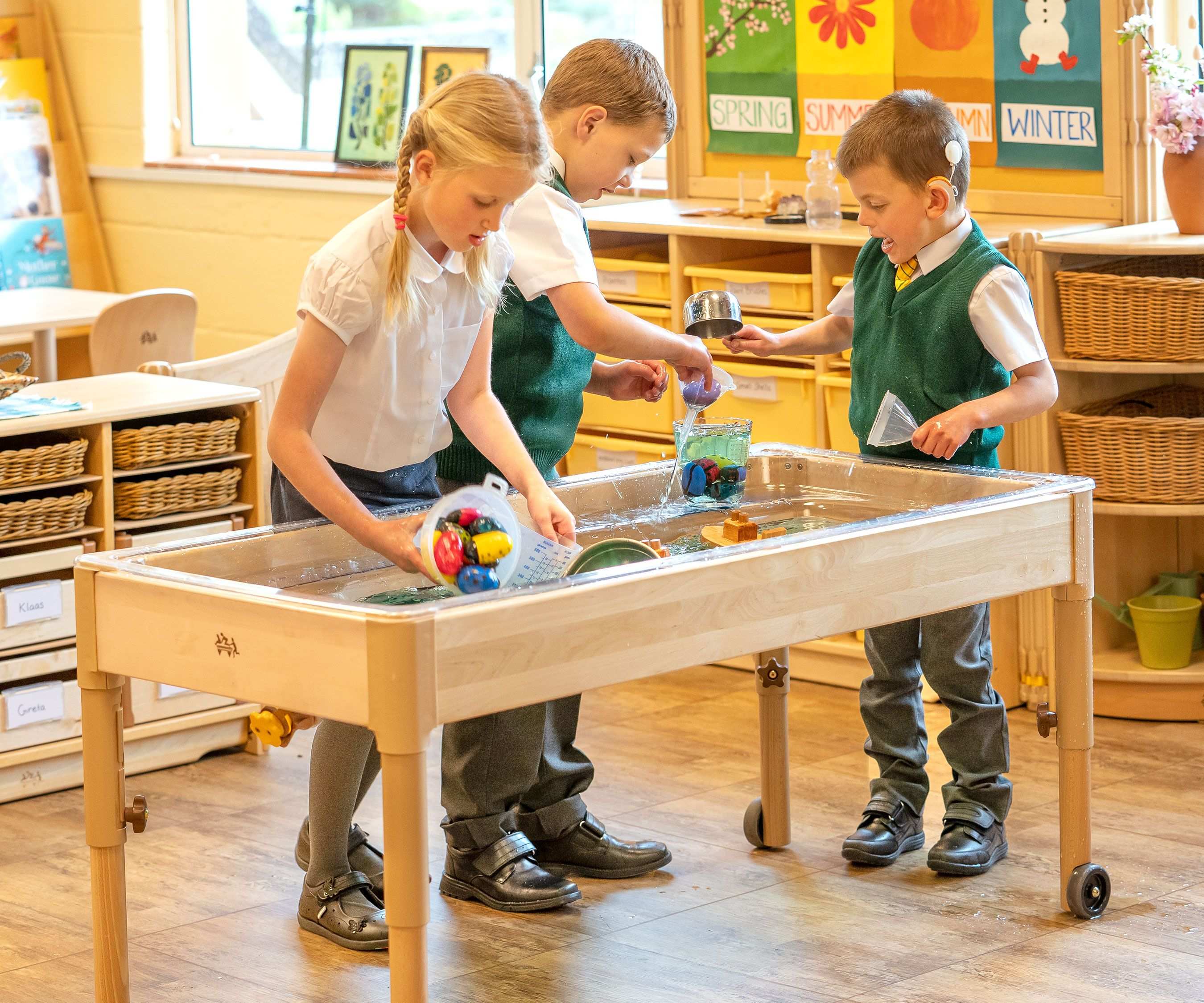 Three children in school uniforms playing with water and toys in a wooden water table.