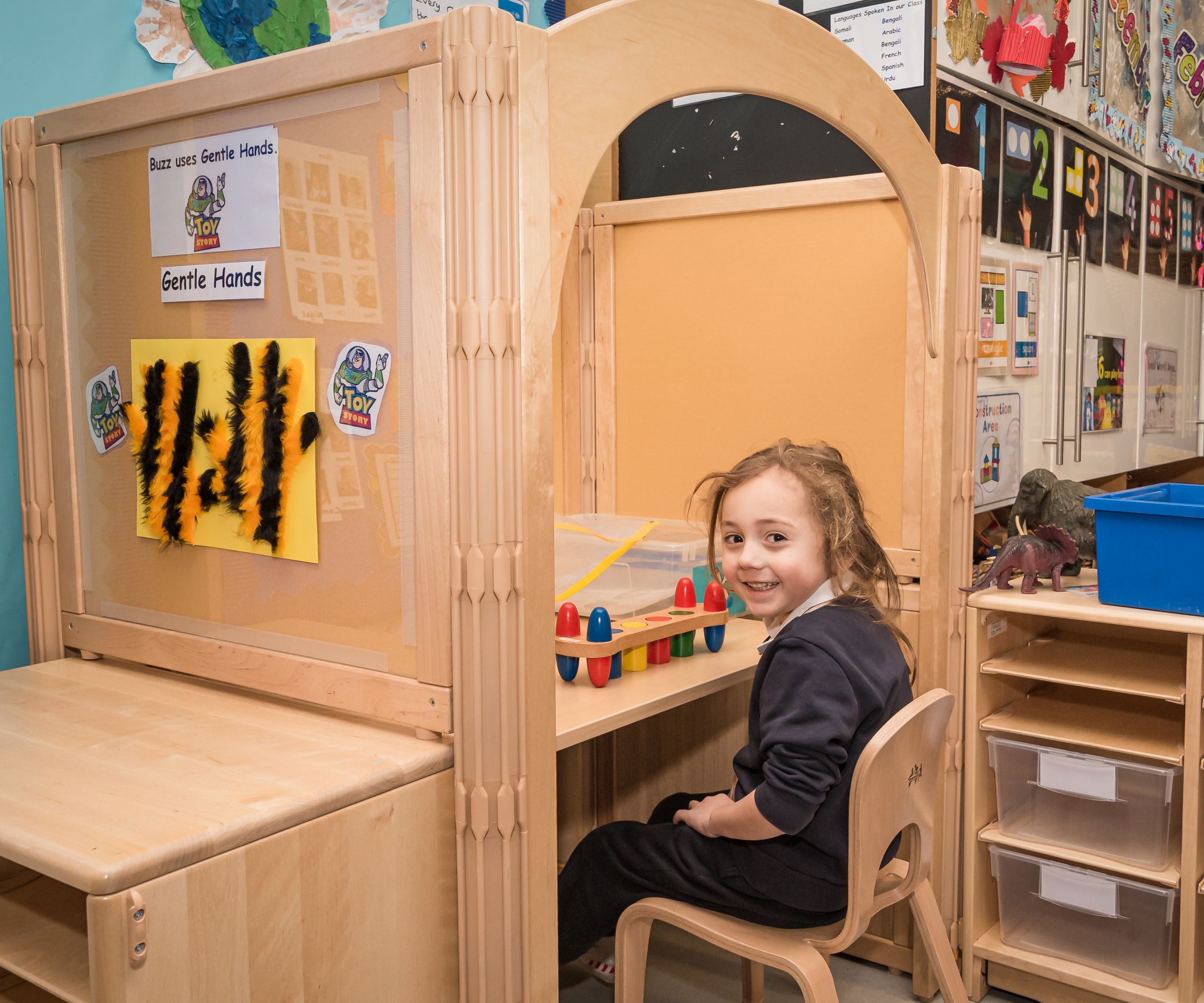 A smiling child sits at a wooden nursery workstation with a desk and storage, playing with a colorful toy.
