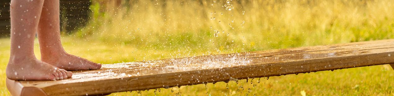 Child's bare feet standing on a wet wooden plank, with water splashing around.