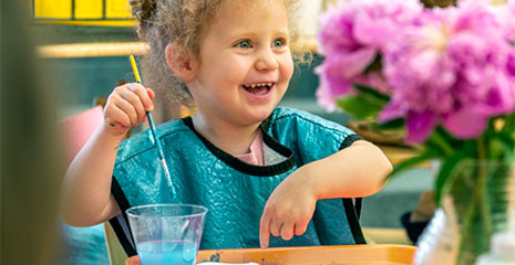 A laughing girl in a blue apron pointing at her painting