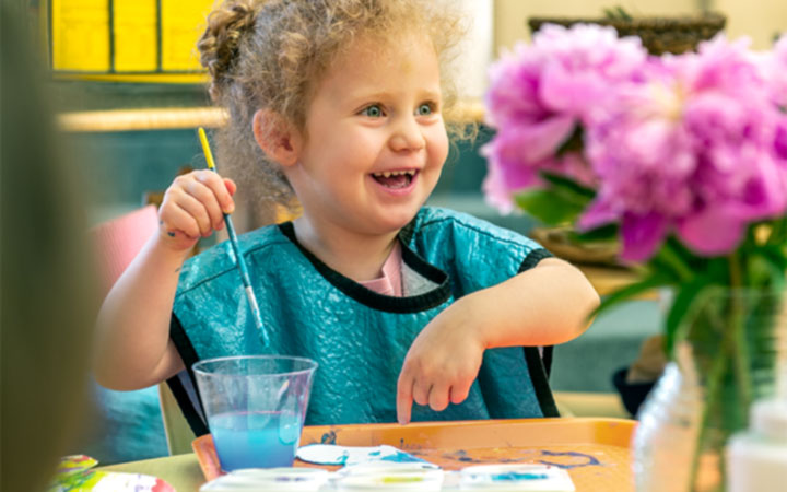 A laughing girl in a blue apron pointing at her painting