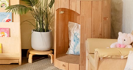 A close-up view of a wooden Hollow Nook with a soft pillow featuring a seahorse design. Next to it is a beige child-sized armchair with a pink plush bear. A potted plant and a wooden bookshelf with children's books complete the inclusive space.