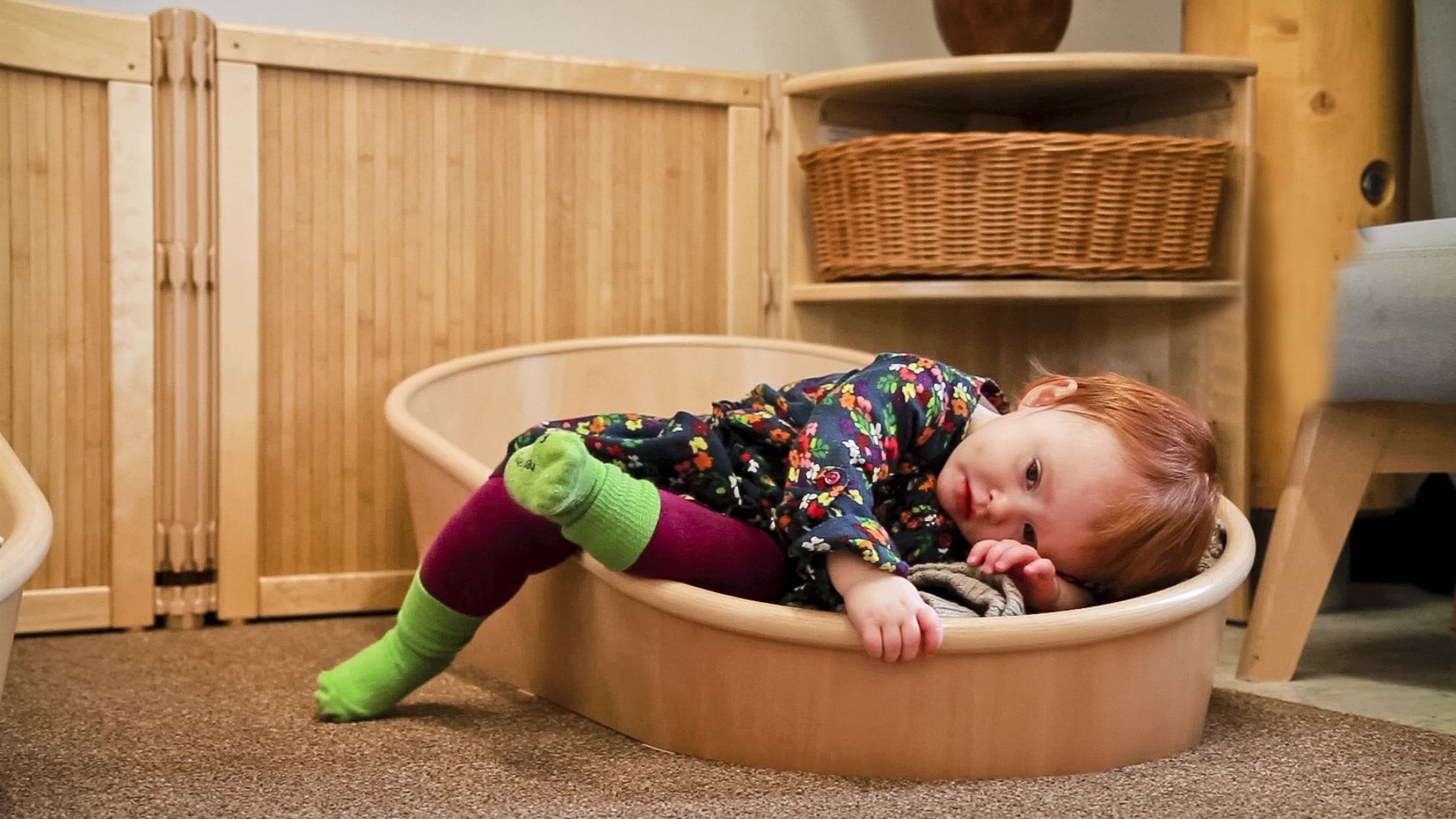 Toddler with red hair and floral dress resting in a wooden Dream Coracle.