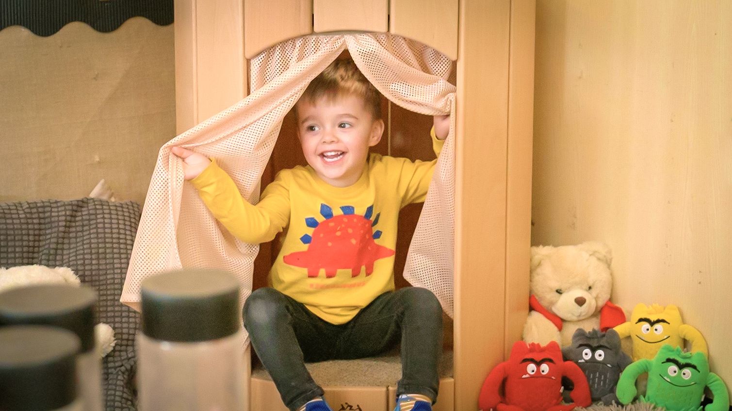 Smiling boy in a yellow dinosaur shirt peeking out from a wooden playhouse with a mesh curtain.