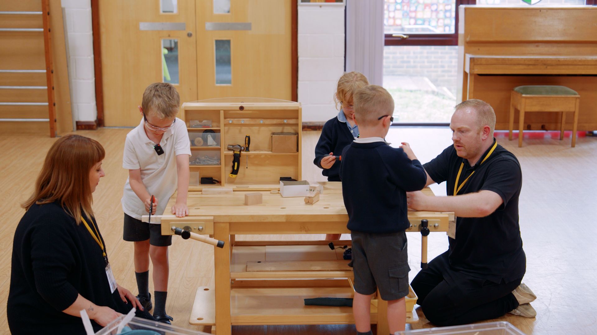 Two adults supervise three children using a wooden workbench with tools and materials in a classroom.