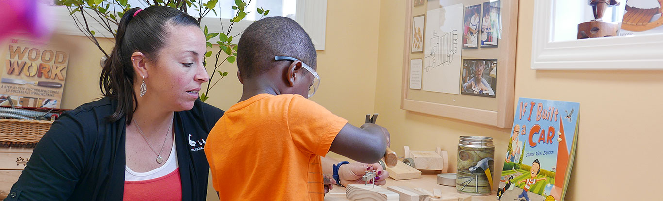 An adult and a child wearing safety glasses work together at a Workbench, hammering nails into wood.