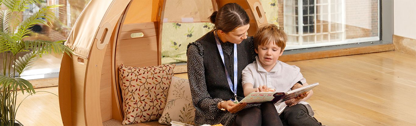 Teacher and child reading a book together inside a Calm Corner, with patterned cushions and a plant nearby.