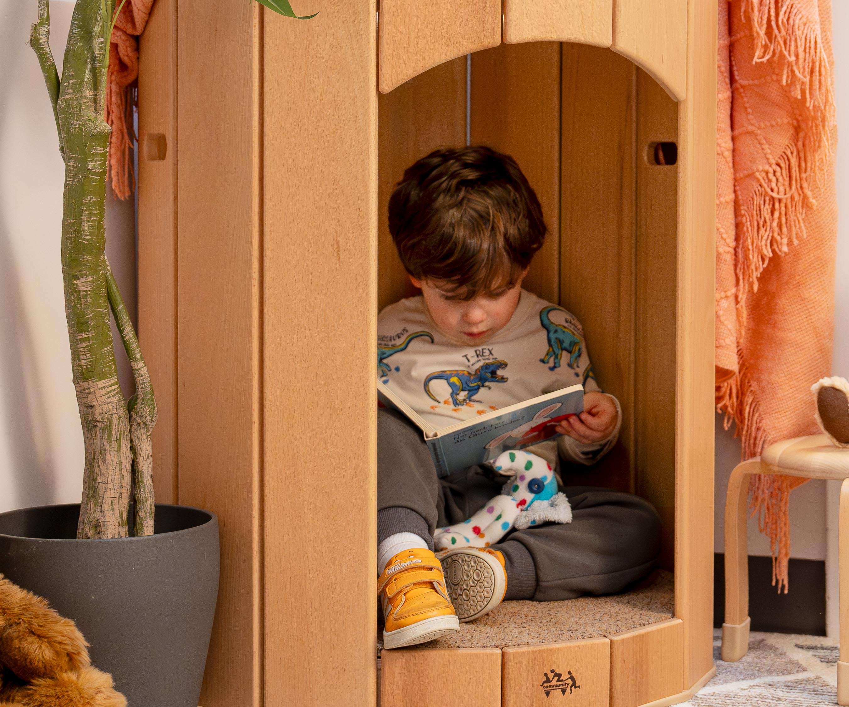 A young child sits inside a wooden Calm Corner reading a book, with a plant and blanket nearby.