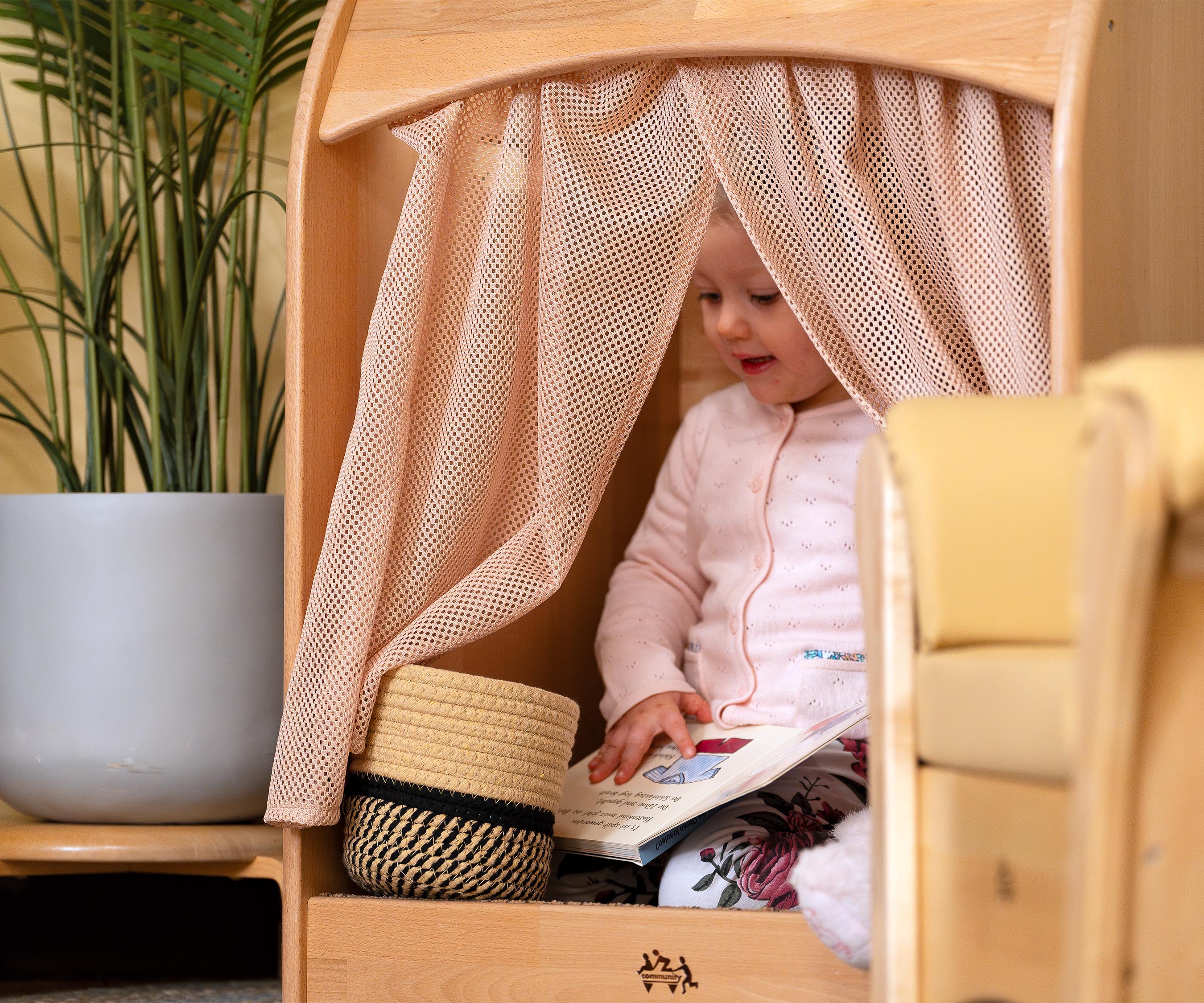 A toddler girl wearing a pink cardigan seen through the curtains of a Nest Nook