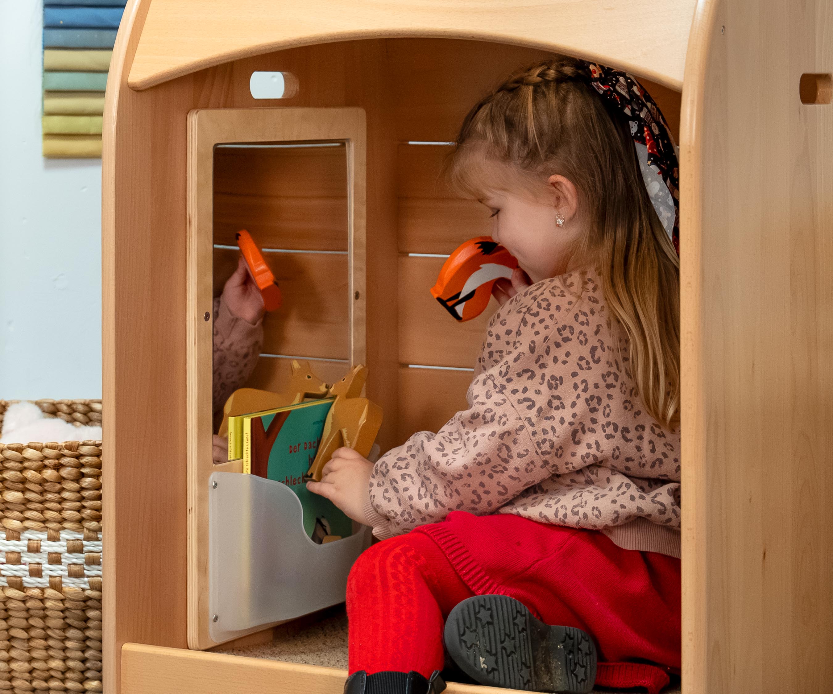 A toddler girl sitting in a Nest Nook calm corner and playing with a wooden fox and looking at books
