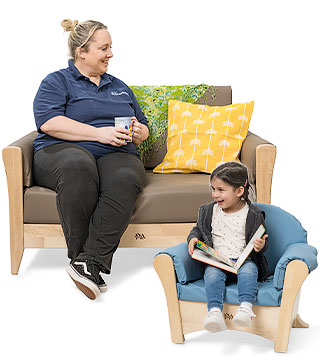 A teacher sitting on a nursery sofa holding a coffee cup and a child sitting in a child's armchair looking at a book.