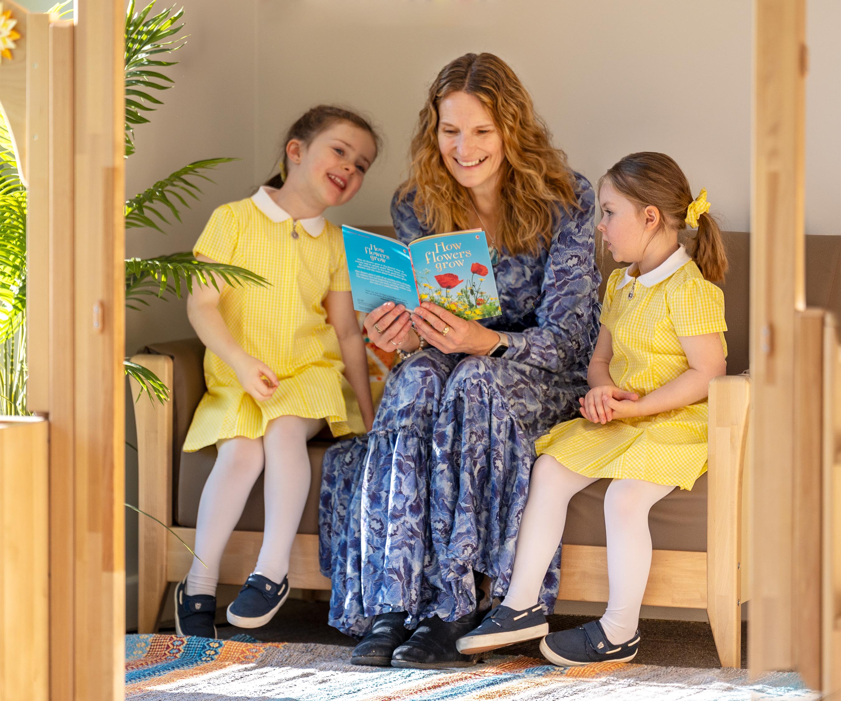A reception teacher and two girls in yellow uniforms reading a story together on a Nursery sofa
