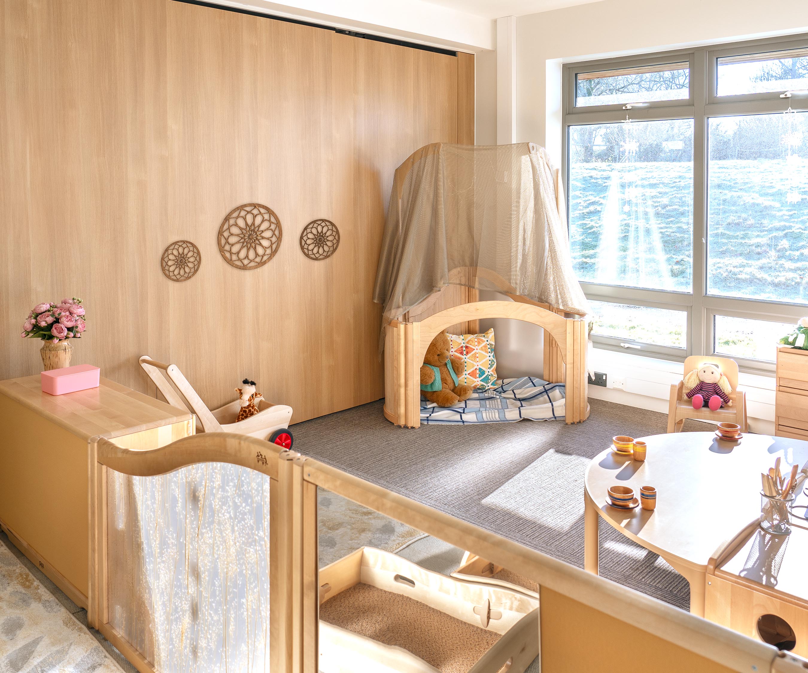 Nursery classroom furnished with solid wood furniture including a cosy den roofed with a sage green mesh fabric.