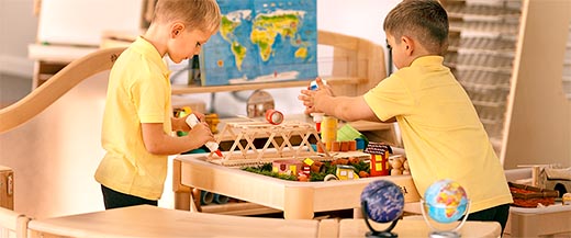 Two children playing with a wooden bridge and small houses on a Roomscapes play table.