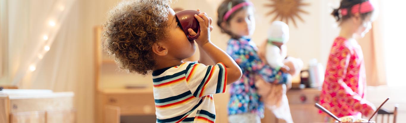 A nursery aged boy in the role play corner pretending to drink tea from a maroon china cup.
