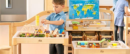 A young boy glues craft materials together on a wooden activity tray table, creating a small village scene.