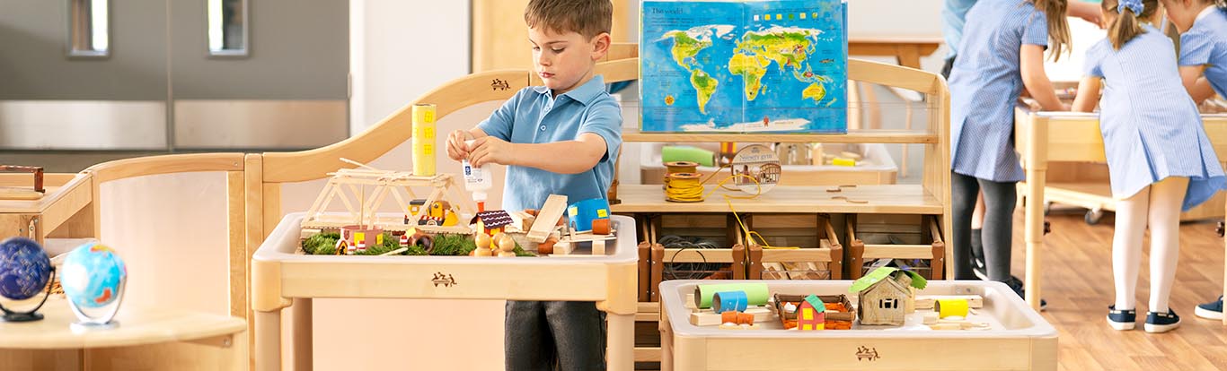 A boy glues a craft project on a tray table, while other children play at similar tables.