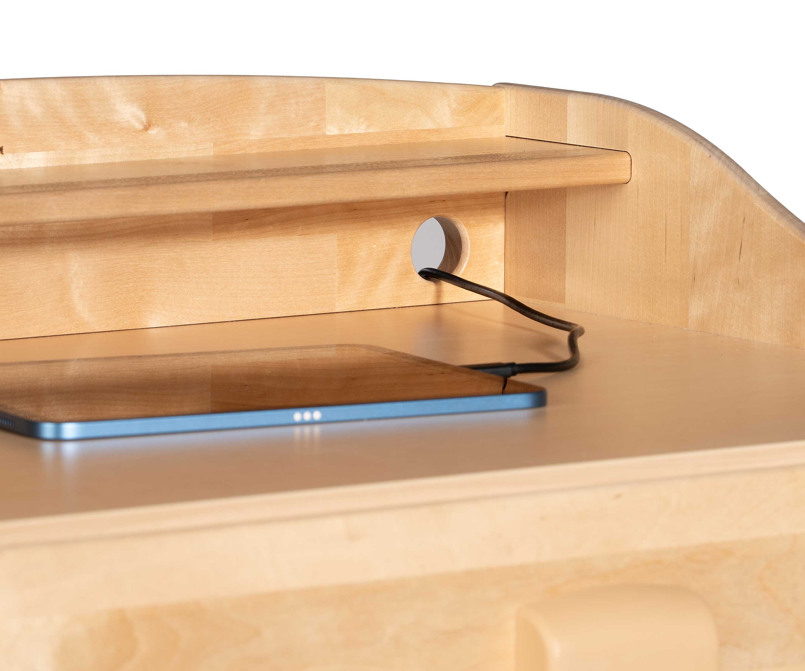 A computer tablet on the writing surface of a teacher standing desk with its charging cable going through one of the cable ports of the desk