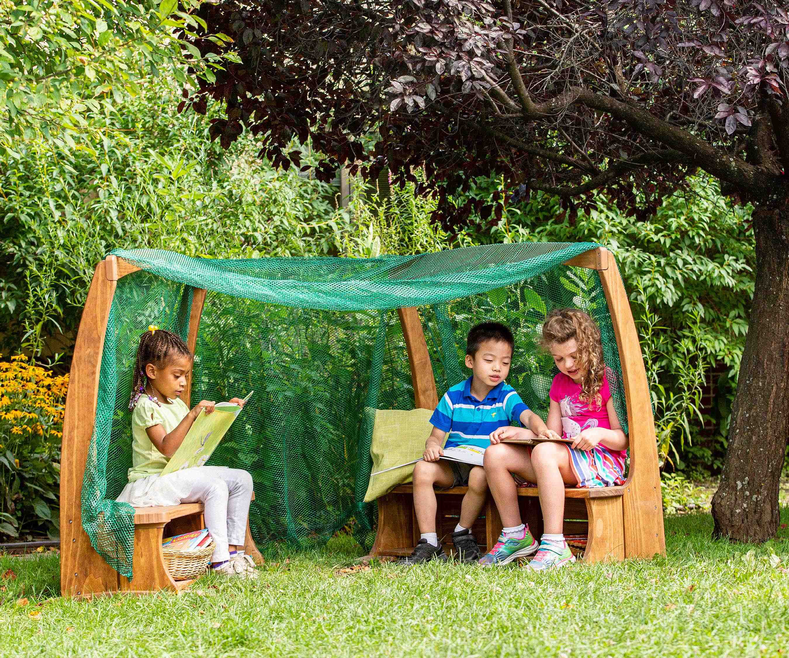Three children reading books inside a wooden Arbour with a green mesh canopy in a garden.