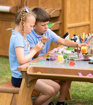 A primary school aged boy in school uniform sitting at a solid wood outdoor table and examining nature objects with a microscope