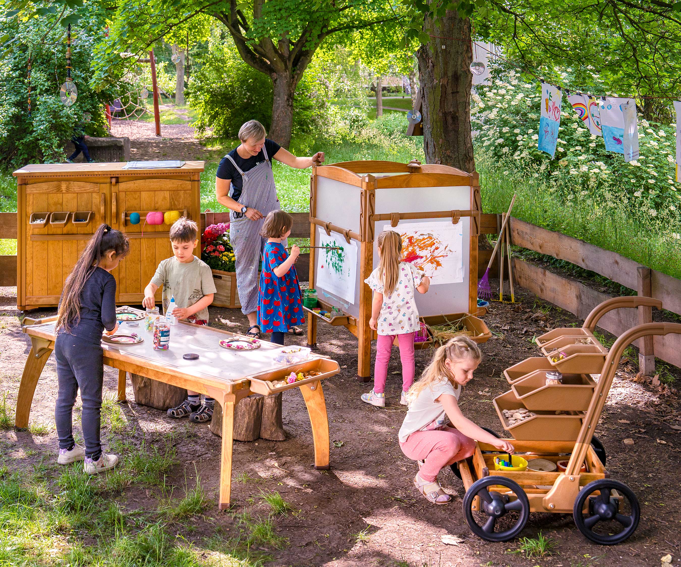 A outdoor classroom for reception and primary school with children engaged in learning at and outdoor table and easel. Materials are stored in an art shed and trolley.
