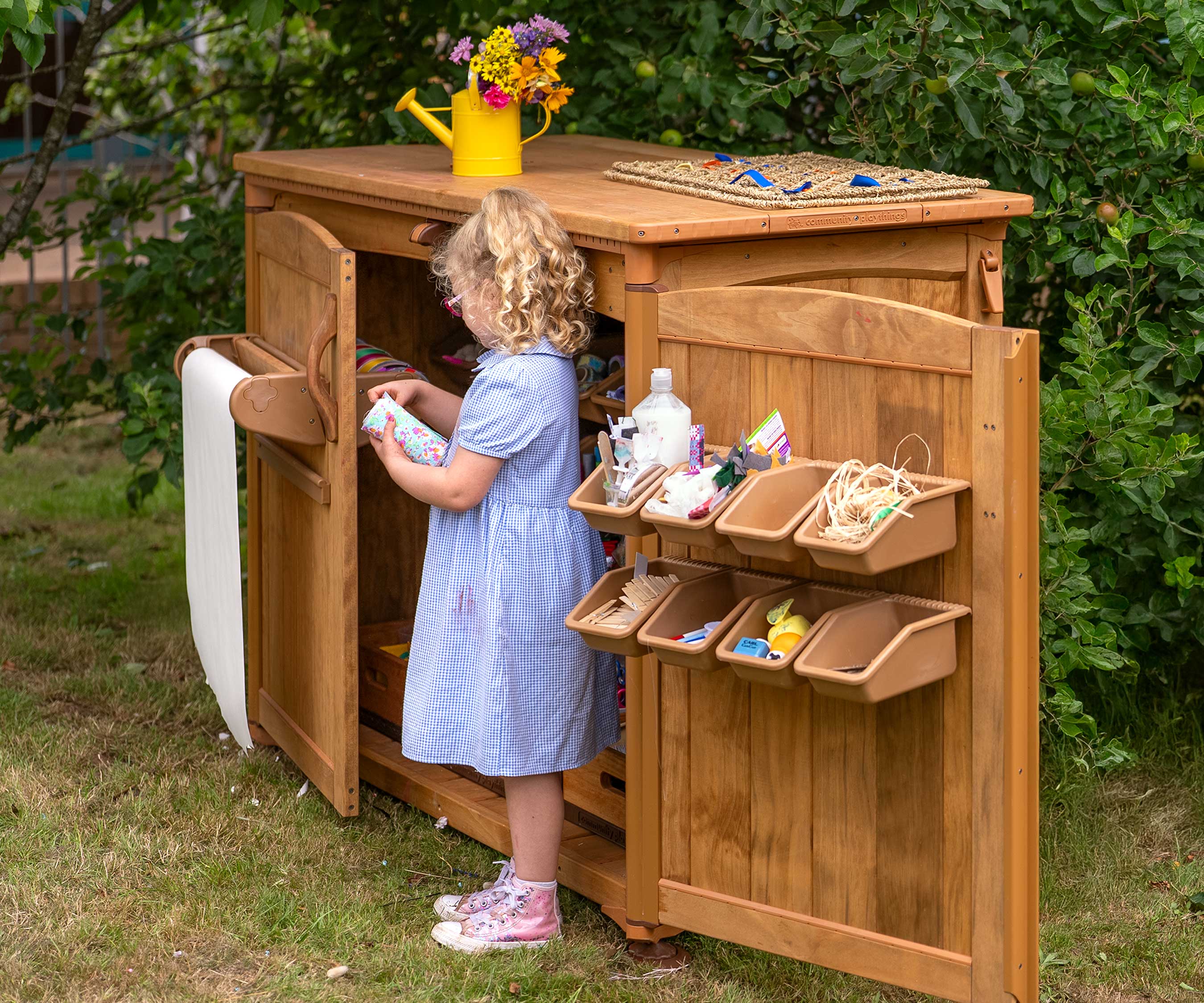 A reception aged child choosing art materials from an outdoor art supply shed. The setting is a primary school outdoor area.
