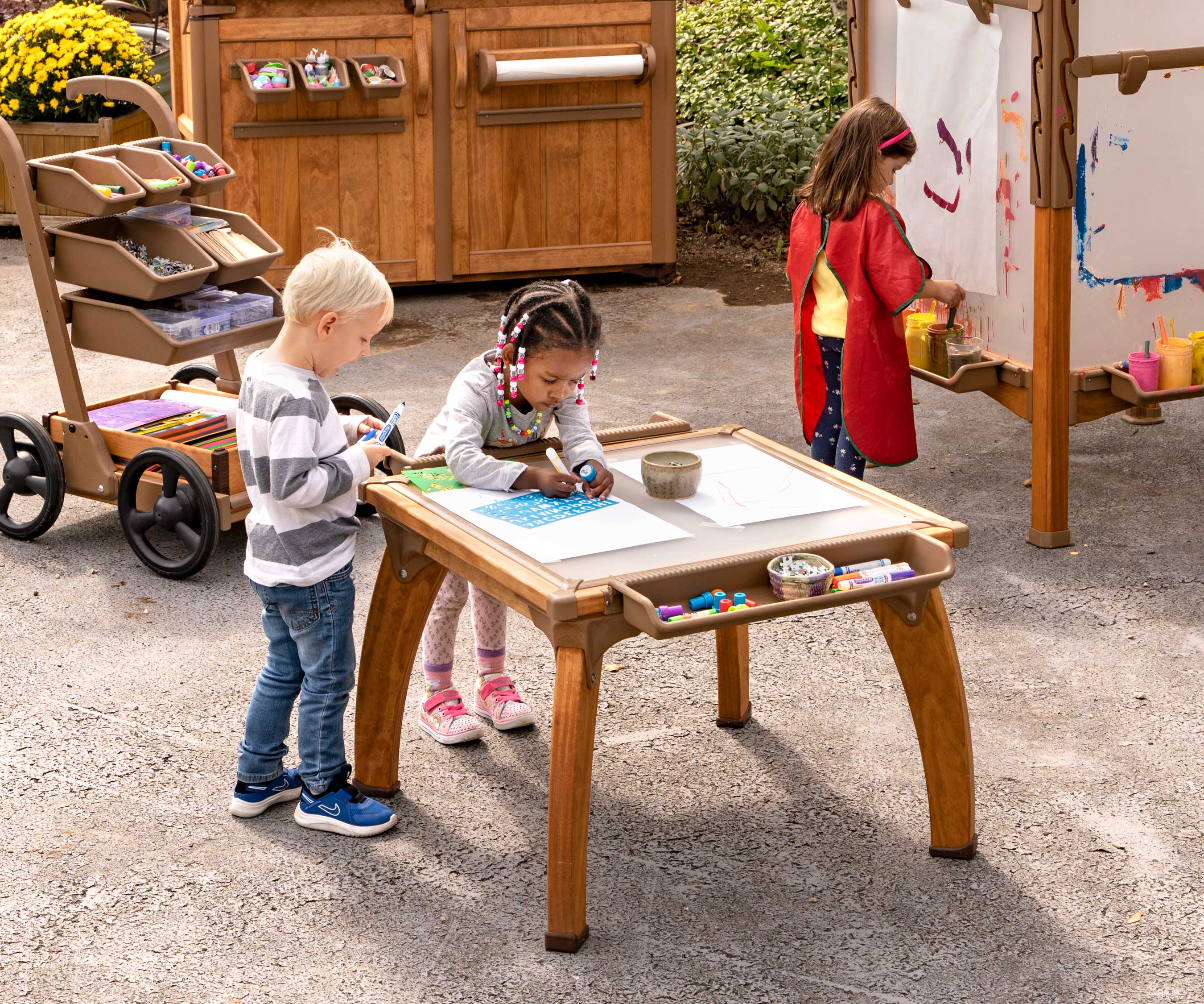 An outdoor art studio with children dawing at a table and a child painting at an easel.