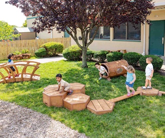 Toddlers playing on Outlast Collection outdoor equipment, including a bridge, tunnel, and stepping stones, on a sunny day.