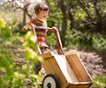 A boy pushing a children’s wooden wheelbarrow through the woods