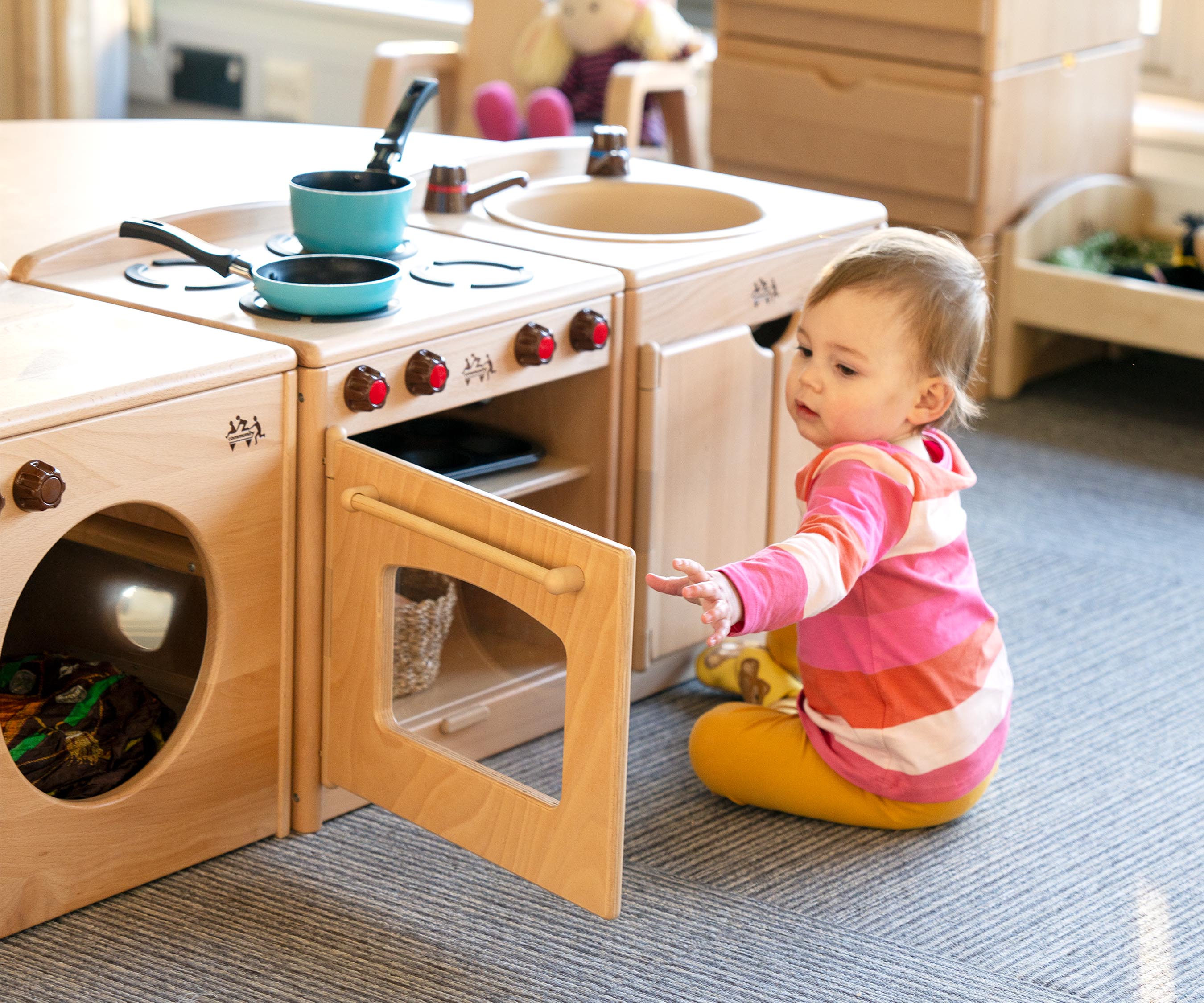 A one year old baby in a striped pink shirt and yellow leggings playing at a solid wood Starter home corner
